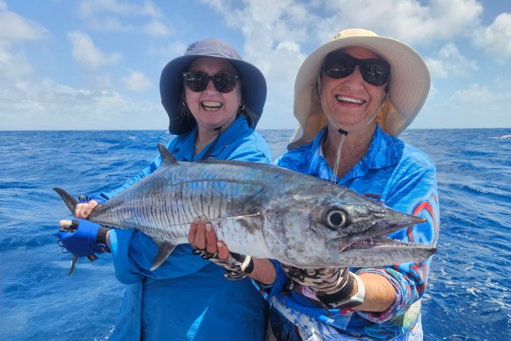 Two people smiling and holding a large fish on a boat in the ocean.
