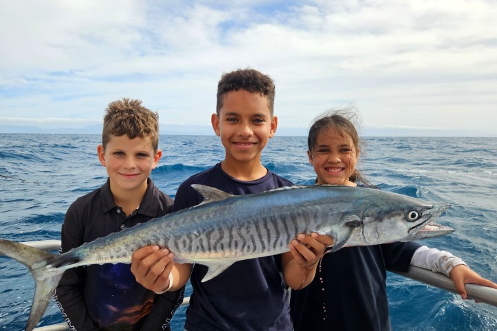 Three children on a boat holding a large fish against a sea backdrop.