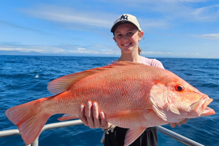 Person smiling, holding a large red fish on a boat with ocean in background.