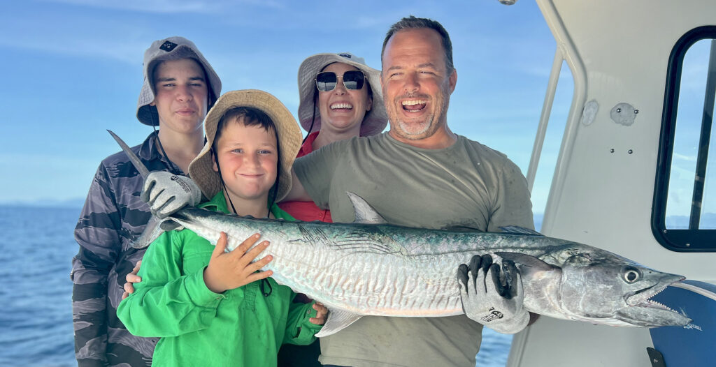 Group on a boat posing with a large fish, smiling, ocean visible in the background.
