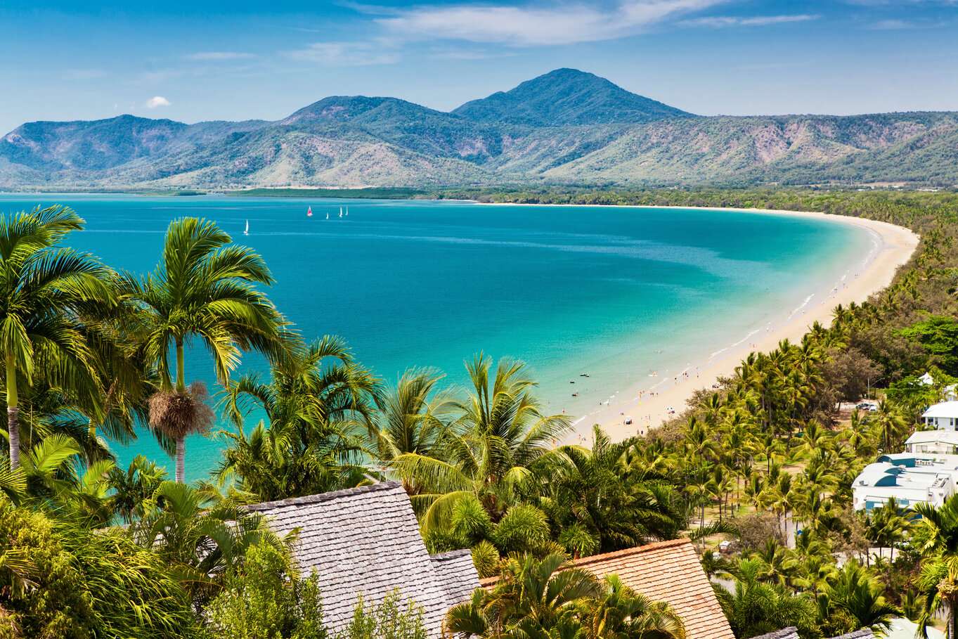 Tropical beach with palm trees, clear water, and distant mountains under a blue sky.